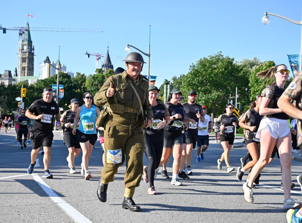 A man in a historical Army uniform gives a thumbs up as he and other runners pass. The Peace Tower can be seen in the background. | Un homme vêtu d'un uniforme militaire historique lève le pouce alors que lui et d'autres coureurs passent devant lui. La Tour de la Paix est visible à l'arrière-plan.
