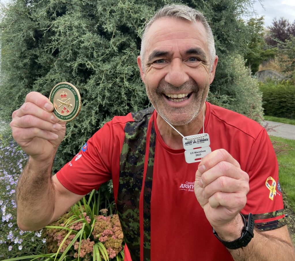 A man smiles and holds up his Canada Army Run coin and medal. | Un homme sourit et brandit sa médaille et sa pièce commémorative de la Course de l'Armée du Canada.